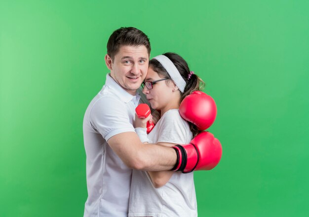 Junger sportlicher Paarmann mit Boxhandschuhen, die seine Freundin glücklich und positiv umarmend über grüner Wand umarmend