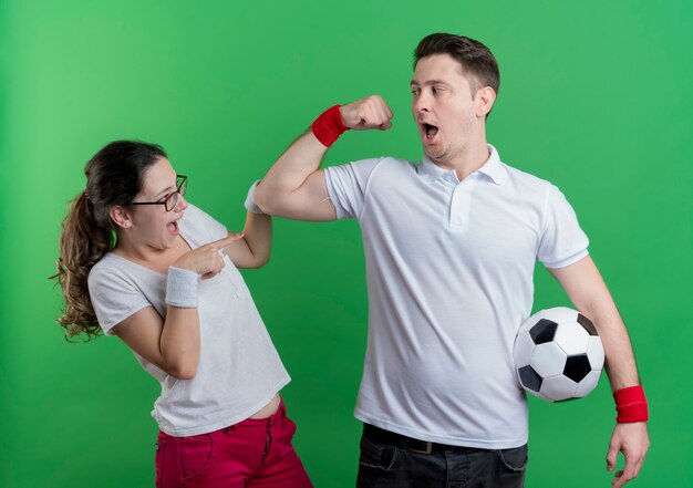Junger sportlicher Paarmann mit Fußball, der Bizeps zu seiner überraschten Freundin zeigt, die über grüner Wand steht