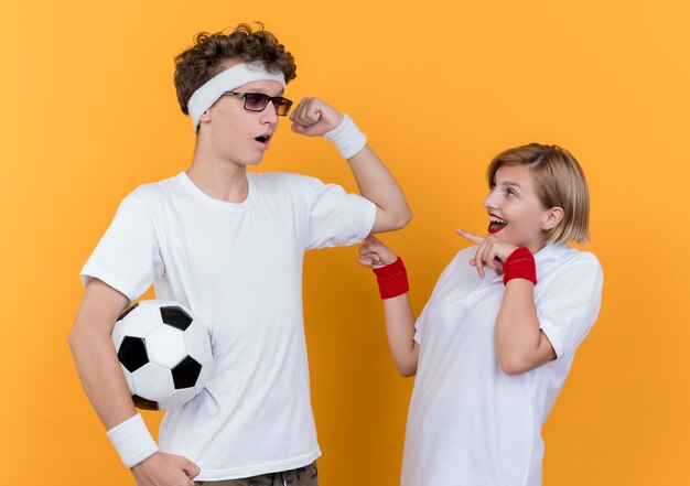 Junger sportlicher Paarmann mit Fußball, der Bizeps zu seiner überraschten Freundin zeigt, die über orange Wand steht