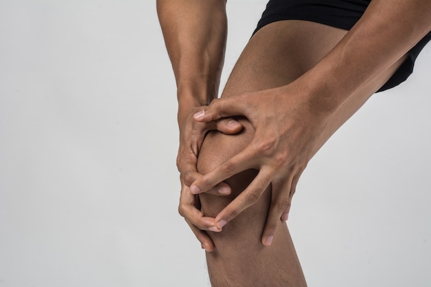 Junger Sportmann mit den starken athletischen Beinen, die Knie mit seinen Händen in den Schmerz halten, nachdem die Ligaturverletzung lokalisiert auf Weiß verletzt worden ist.