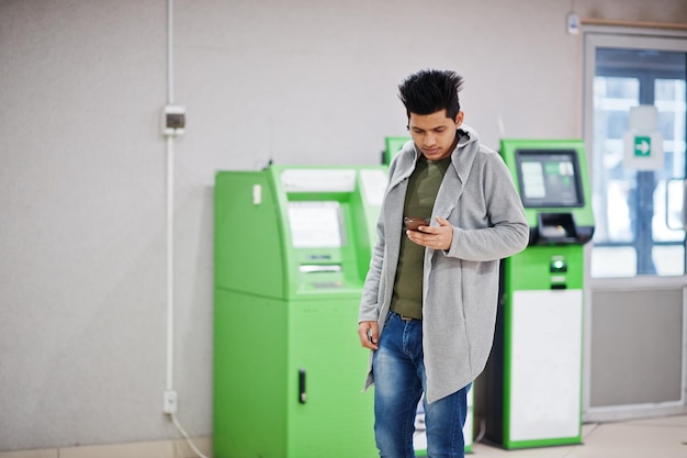 Junger stilvoller asiatischer Mann mit Handy gegen Reihe des grünen Geldautomaten