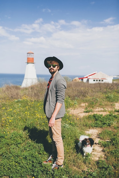 Junger stilvoller Hipster-Mann im Hut, der mit Hund in der Landschaft geht
