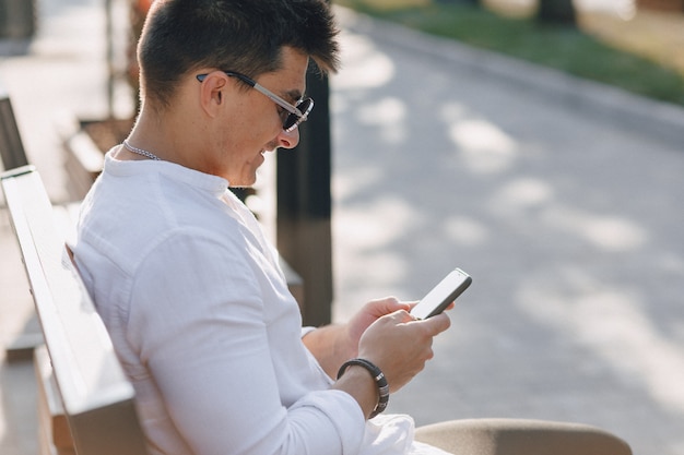 Junger stilvoller Kerl im Hemd mit Telefon auf Bank am sonnigen warmen Tag draußen