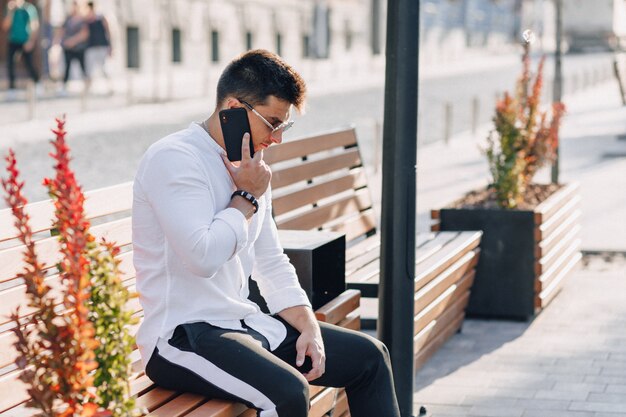 Junger stilvoller Kerl im Hemd mit Telefon auf Bank am sonnigen warmen Tag draußen