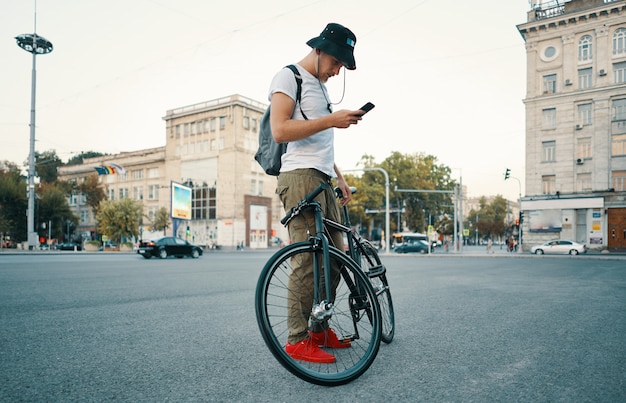 Junger stilvoller Mann, der am Telefon eine SMS schreibt, während er Fahrrad beiseite stellt.