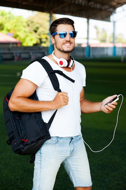 Junger stilvoller Mann, der auf einem Fußballfeld aufwirft, hören Musik. Kopfhörer auf seinen Schultern, Spieler in der Hand, Sonnenbrille im Gesicht