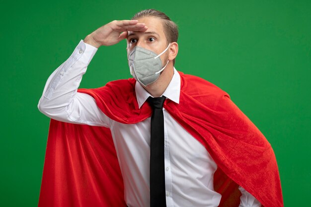 Junger Superheldenmann, der medizinische Maske und Krawatte trägt, die Entfernung mit Hand lokalisiert auf Grün betrachtet