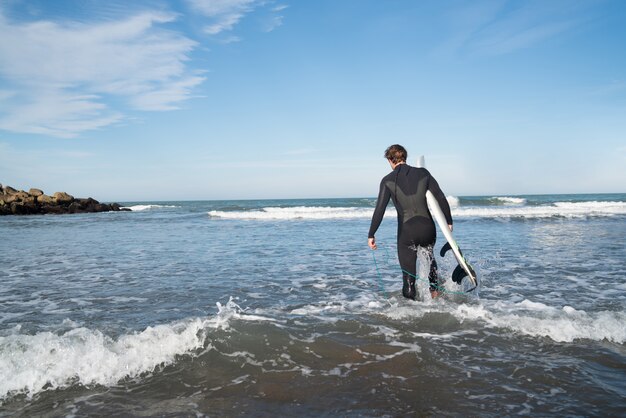 Junger Surfer, der mit seinem Surfbrett in einem schwarzen Surfanzug ins Wasser tritt. Sport- und Wassersportkonzept.