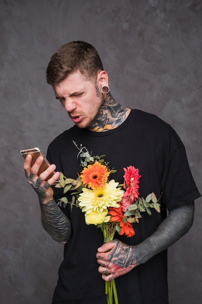 Kostenloses Foto junger tätowierter und durchbohrter mann, der in der hand den blumenblumenstrauß macht bei der anwendung des handys hält