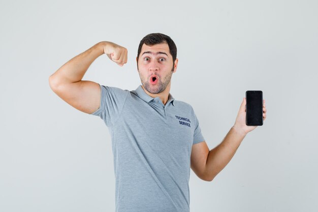 Junger Techniker in der grauen Uniform, die Handy hält, Muskeln zeigt und zuversichtlich schaut, Vorderansicht.