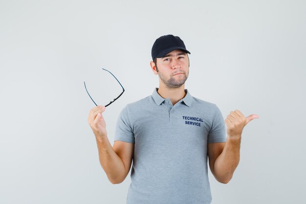 Junger Techniker in grauer Uniform, der mit Daumen zeigt, Brille hält und zögernd aussieht.