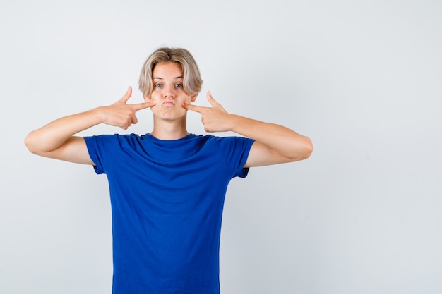 Junger Teenager, der auf seine geschwollenen Wangen im blauen T-Shirt zeigt und verwirrt aussieht. Vorderansicht.