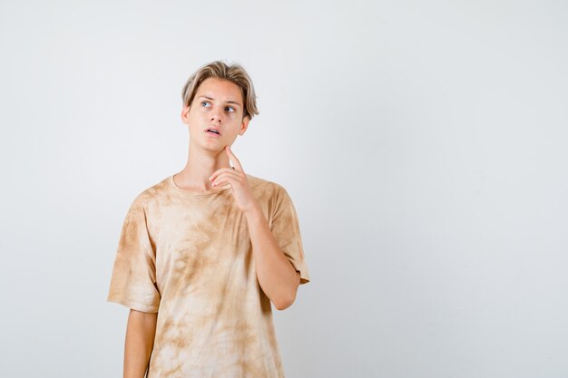 Junger Teenager, der den Finger am Kiefer hält, im T-Shirt nach oben schaut und nachdenklich aussieht, Vorderansicht.