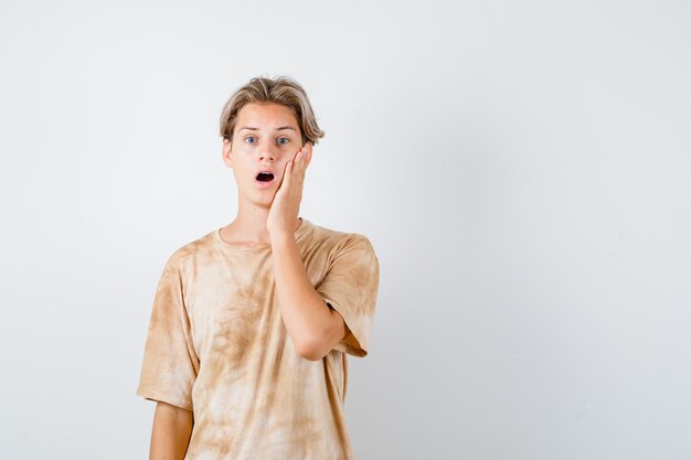 Junger Teenager, der die Hand auf der Wange im T-Shirt hält und überrascht, Vorderansicht schaut.