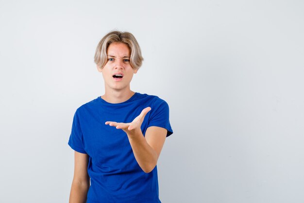 Junger Teenager, der die Hand vorne im blauen T-Shirt ausdehnt und verwirrt aussieht. Vorderansicht.