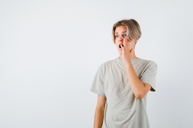 Junger Teenager im T-Shirt, der die Hand in der Nähe des offenen Mundes hält, während er wegschaut und schockiert aussieht, Vorderansicht.