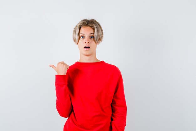 Junger Teenagerjunge im roten Pullover, der mit dem Daumen nach links zeigt und überrascht, Vorderansicht schaut.