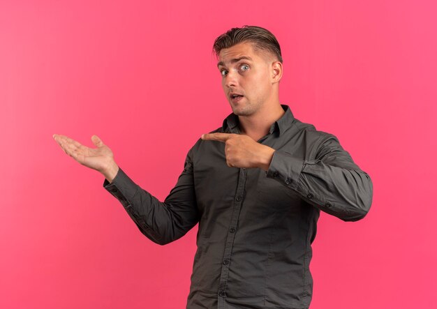Junger überraschter blonder hübscher Mann zeigt auf leere Hand lokalisiert auf rosa Hintergrund mit Kopienraum