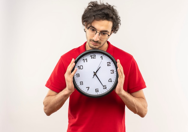 Junger überraschter Mann im roten Hemd mit optischer Brille hält und schaut auf Uhr lokalisiert auf weißer Wand