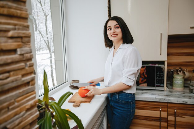 junger und hübscher Brunette, der das Fenster in der Küche mit Orange bereitsteht