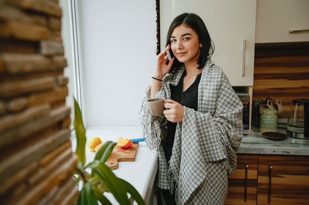 junger und hübscher Brunette, der in der Küche mit Telefon steht und einen Kaffee trinkt