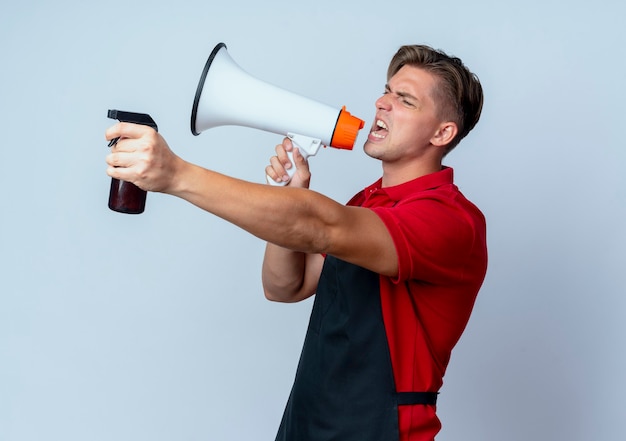 Junger verärgerter blonder männlicher Friseur in der Uniform hält Lautsprecher und Sprühflasche lokalisiert auf weißem Hintergrund mit Kopienraum