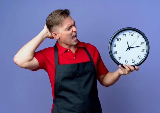 Junger verärgerter blonder männlicher Friseur in Uniform legt Hand hinter Kopf, der Uhr hält und auf Uhr lokalisiert auf violettem Raum mit Kopienraum betrachtet