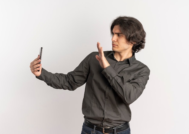 Junger verärgerter hübscher kaukasischer Mann hält und betrachtet Telefon mit erhabener Hand lokalisiert auf weißem Hintergrund mit Kopienraum