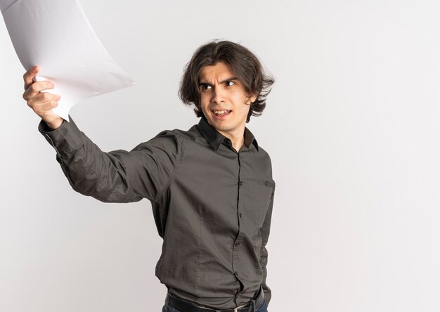 Junger verärgerter hübscher kaukasischer Mann hebt leere weiße Papierblätter auf und schaut auf Seite lokalisiert auf weißem Hintergrund mit Kopienraum