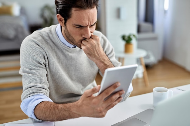 Junger verstörter Geschäftsmann, der zu Hause arbeitet und problematische E-Mails auf einem digitalen Tablet liest