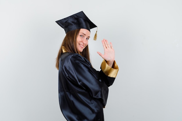Junger weiblicher Absolvent, der Hand für Begrüßung im akademischen Kleid und in der fröhlichen Vorderansicht winkend wendet.