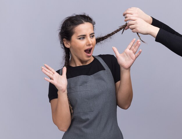 Junger weiblicher Friseur in der Uniform jemand, der ihr Haar isoliert auf weißer Wand schneidet