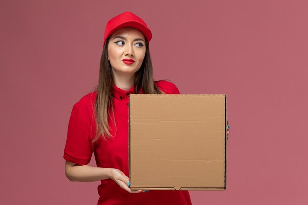 Junger weiblicher kurier der vorderansicht in der roten uniform, die lieferung-nahrungsmittelbox auf der rosa schreibtischdienstlieferuniformfirma hält