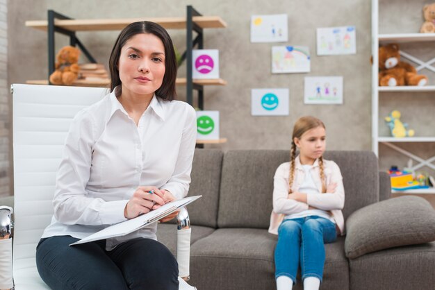 Junger weiblicher Psychologe, der auf Stuhl mit dem Klemmbrett sitzen und der Stift, der vor deprimiertem Mädchen sitzt