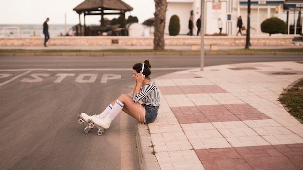Junger weiblicher Schlittschuhläufer, der auf hörender Musik des Bürgersteigs auf Kopfhörer sitzt