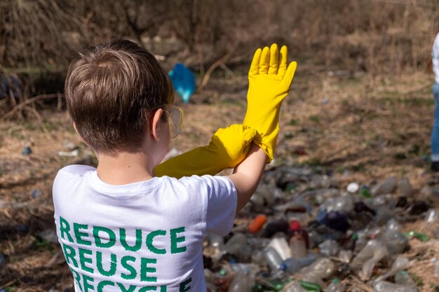 Junger weißer kaukasischer Junge mit Recycling-Symbol auf seinem T-Shirt und Brille, der gelbe Handschuhe anzieht