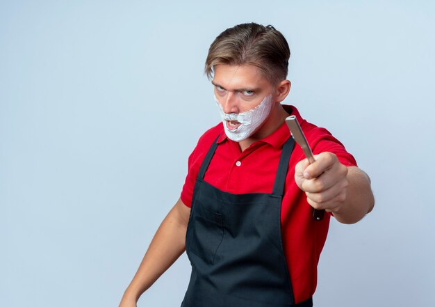 Junger wütender blonder männlicher Friseur im einheitlich verschmierten Gesicht mit Rasierschaum, der gerade Rasiermesser lokalisiert auf weißem Hintergrund mit Kopienraum hält