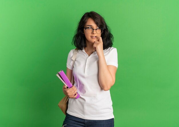 Junges ängstliches hübsches kaukasisches Schulmädchen, das Brille und Rückentasche trägt, beißt Finger, der Bücher auf Grün mit Kopienraum hält