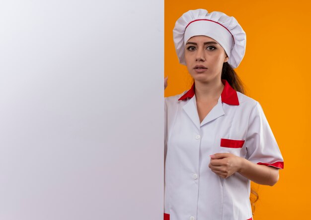 Junges ängstliches kaukasisches Kochmädchen in der Kochuniform steht hinter weißer Wand lokalisiert auf orange Hintergrund mit Kopienraum