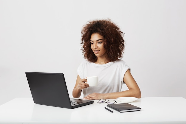 Junges afrikanisches Mädchen, das Tasse hält Laptop betrachtet, der über weiße Wand lächelt.