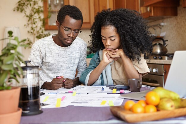 Junges afrikanisches Paar, das Papierkram zusammen macht und am Küchentisch mit vielen Papieren sitzt