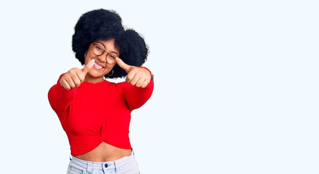 Junges afroamerikanisches Mädchen in lässiger Kleidung und Brille macht zustimmend eine positive Geste mit der Hand, streckt den Daumen nach oben, lächelt und freut sich über den Erfolg. Siegergeste.