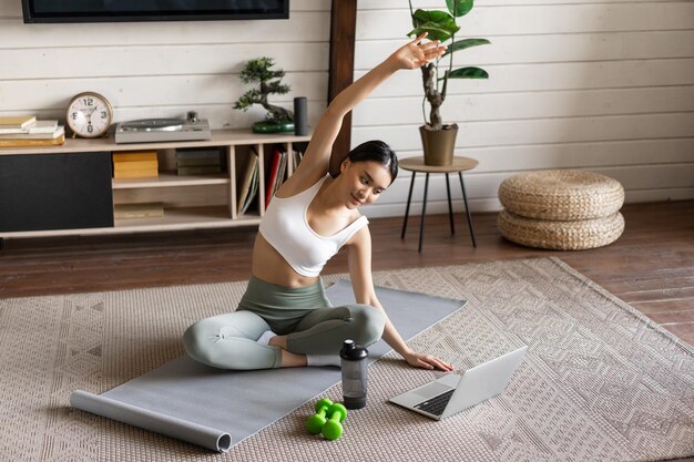 Junges asiatisches Fitnessmädchen, das zu Hause Dehnübungen macht, Arme strecken und auf Laptop f...