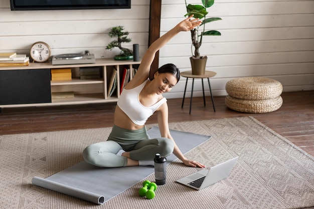 Junges asiatisches Fitnessmädchen, das zu Hause Dehnübungen macht, Arme strecken und auf Laptop f...