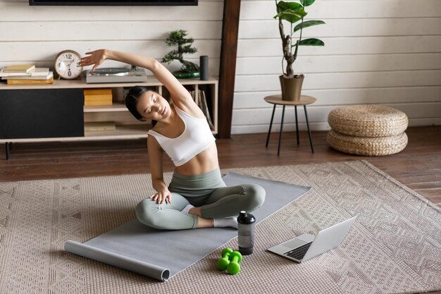 Junges asiatisches Fitnessmädchen, das zu Hause Stretching-Sporttraining macht, die Arme streckt und auf den Laptop schaut ...