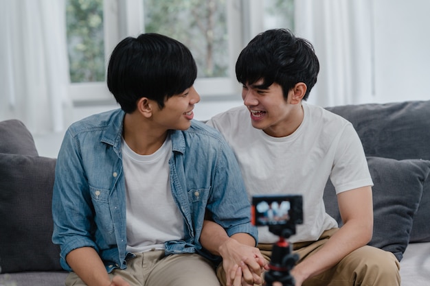 Junges asiatisches homosexuelles Paarinfluencer-Paar vlog zu Hause. Die jugendlich koreanischen glücklichen LGBTQ-Männer entspannen sich Spaß unter Verwendung des Kameraaufzeichnungs-vlog-Video-Uploads in Social Media beim Lügensofa im Wohnzimmer am Haus.