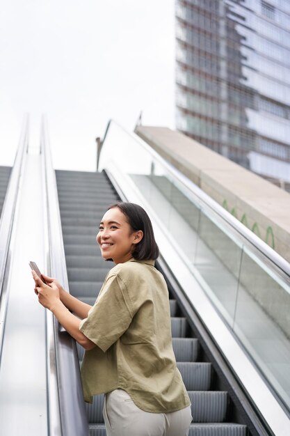Junges asiatisches Mädchen, das auf einer Rolltreppe hochgeht und ein Smartphone lächelnd hält, während es in der Stadt spazieren geht