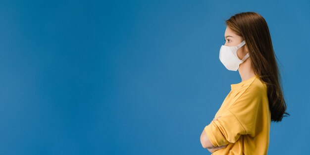 Junges asiatisches Mädchen, das medizinische Gesichtsmaske mit gekleidet in lässigem Stoff und Blick auf Leerzeichen lokalisiert auf blauem Hintergrund trägt. Soziale Distanzierung, Quarantäne für Koronavirus. Panorama Banner Hintergrund.