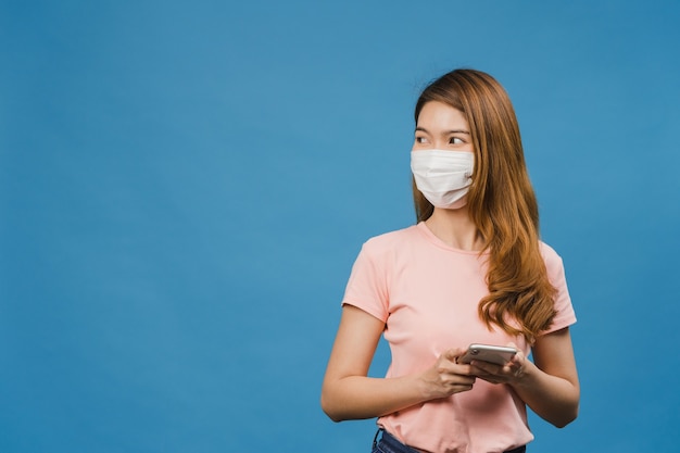 Junges asiatisches Mädchen, das medizinische Gesichtsmaske mit Handy trägt und in Freizeitkleidung gekleidet ist, die auf blauer Wand isoliert ist?