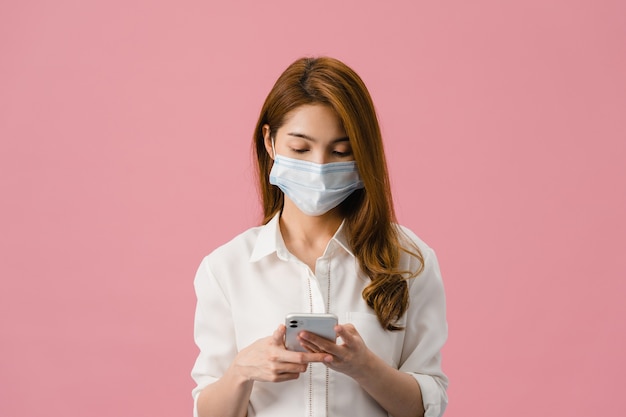 Junges asiatisches Mädchen, das medizinische Gesichtsmaske mit Mobiltelefon trägt und in Freizeitkleidung auf rosafarbenem Hintergrund gekleidet ist.